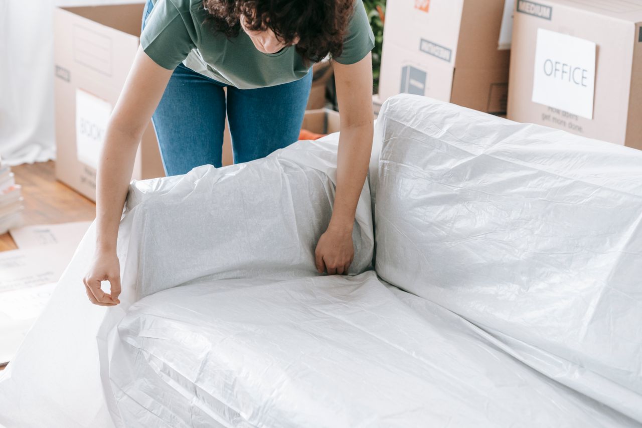 Une femme emballe avec soin un canapé dans un matériau de protection, tandis qu'elle prépare des cartons dans une pièce lumineuse.